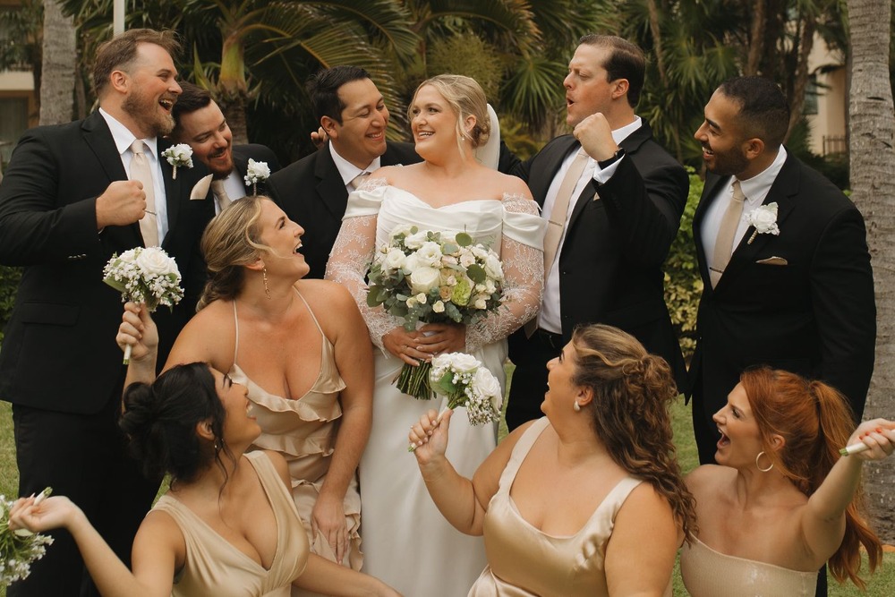 Catherine and Carlos at their destination wedding: Bride in white with bouquet, smiling bridal party outdoors.
