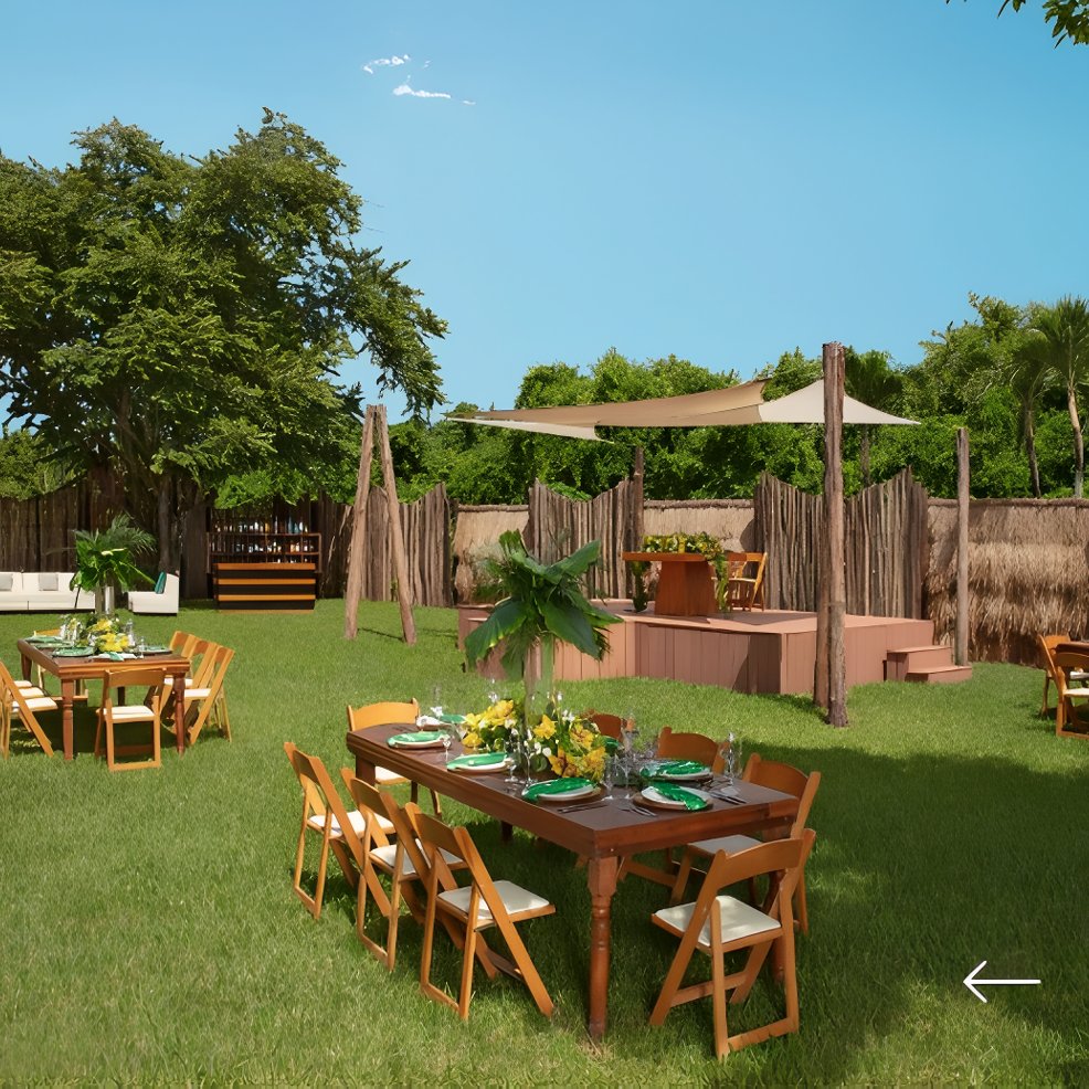 Destination wedding setup with wooden tables and chairs on grass, stage and canopy in the background.