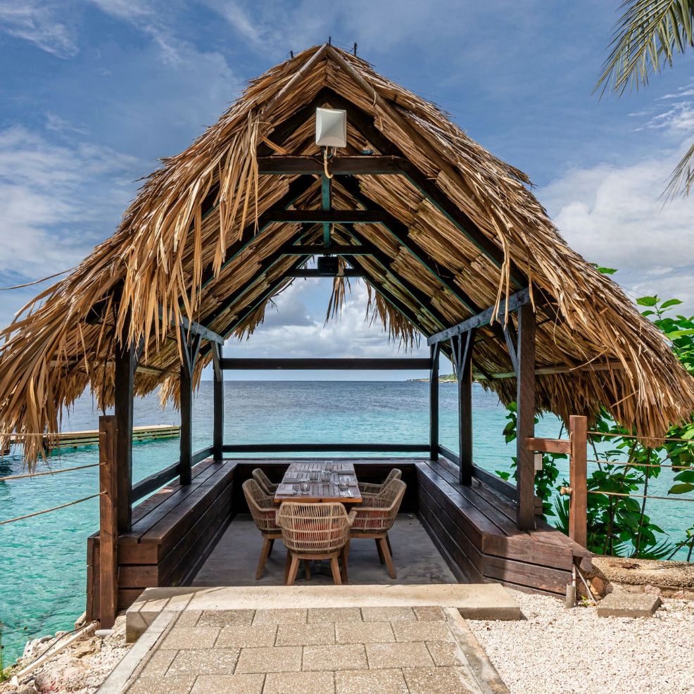A thatched-roof gazebo with a table and chairs overlooks clear blue ocean water, perfect for a wedding.