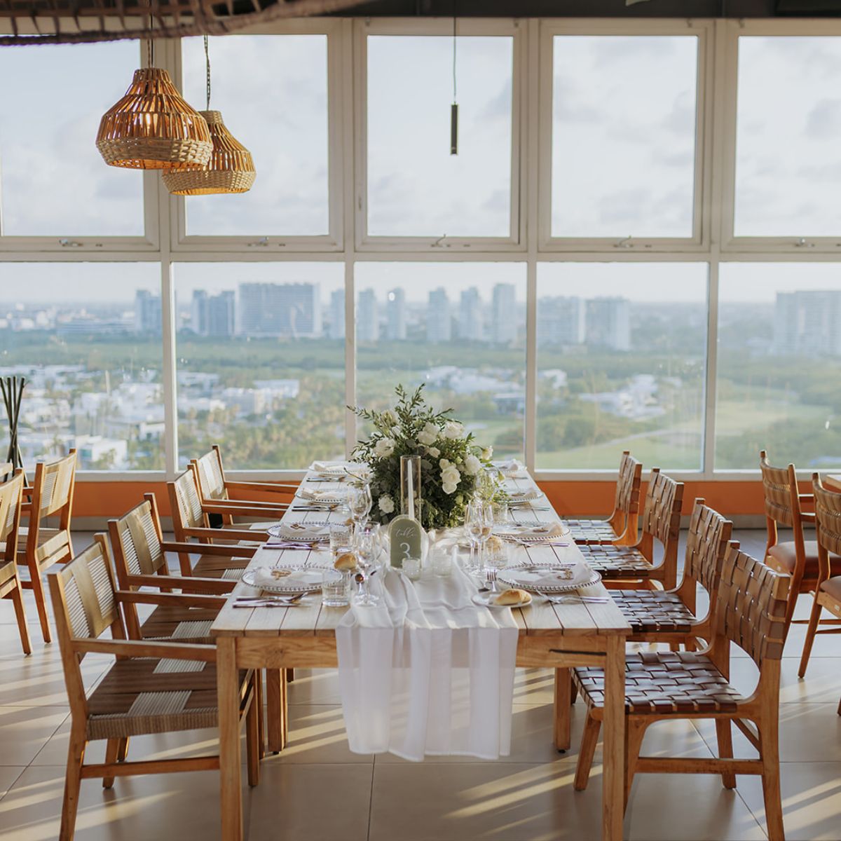 A dining table elegantly set for a destination wedding in a bright room with large windows and city views.