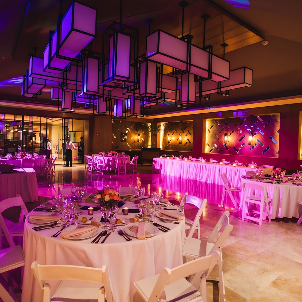 A wedding banquet hall featuring round tables, white chairs, and elegant purple ambient lighting.