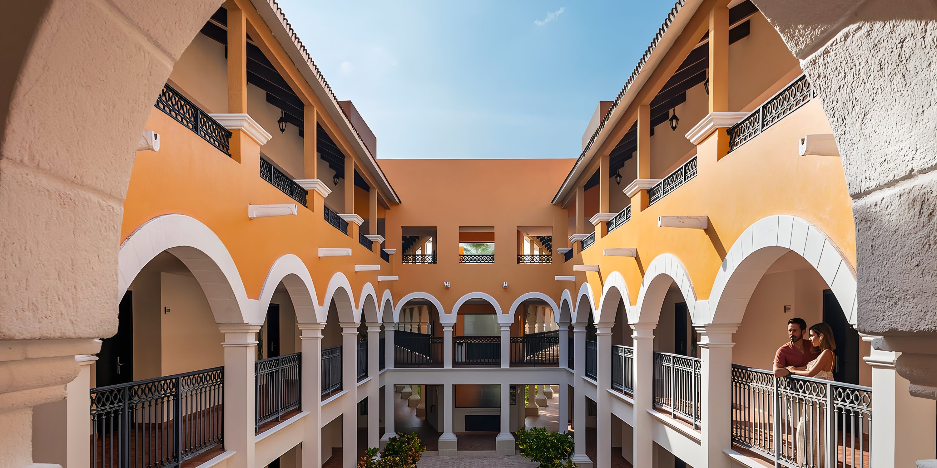 Ocean Coral and Turquesa: Yellow and white arched courtyard with balconies, ideal for destination wedding; two people visible on right.