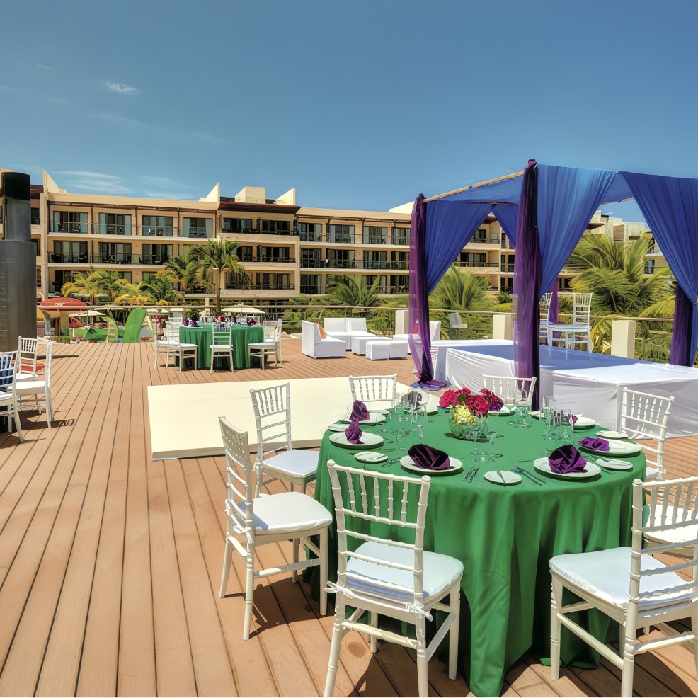 Destination wedding setup outdoors with green tablecloths, white chairs, and purple drapery by a hotel.