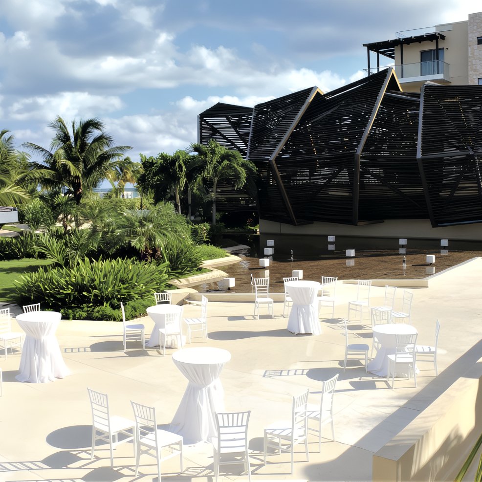 Outdoor patio arranged for a destination wedding, featuring round tables and white chairs by a modern building.