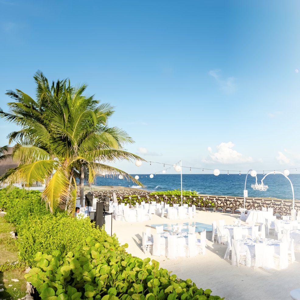 Destination wedding setup with elegant white tables and chairs by the ocean under a clear blue sky.