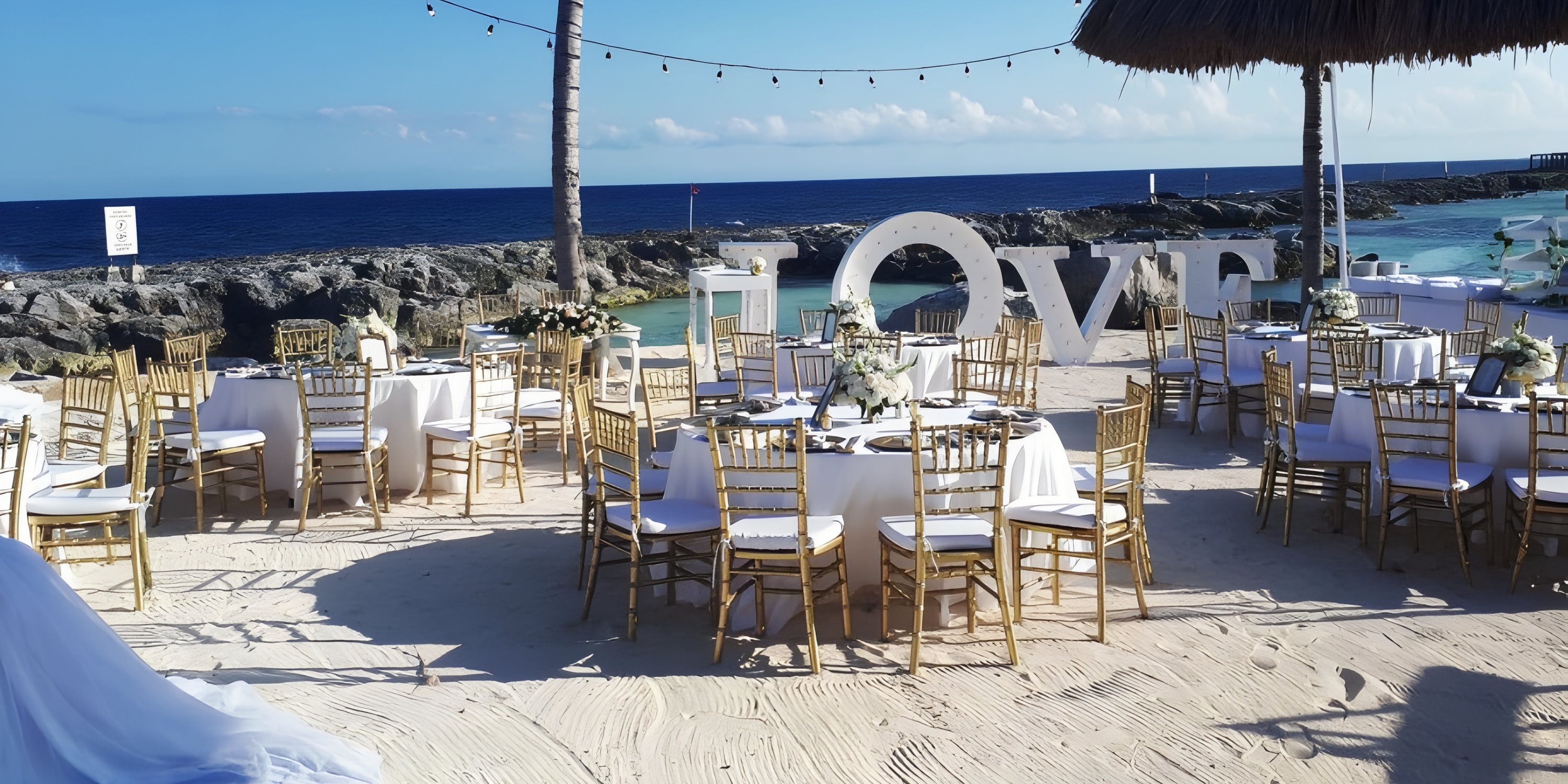 Destination wedding reception on the beach with white tables, gold chairs, and a glowing LOVE sign.