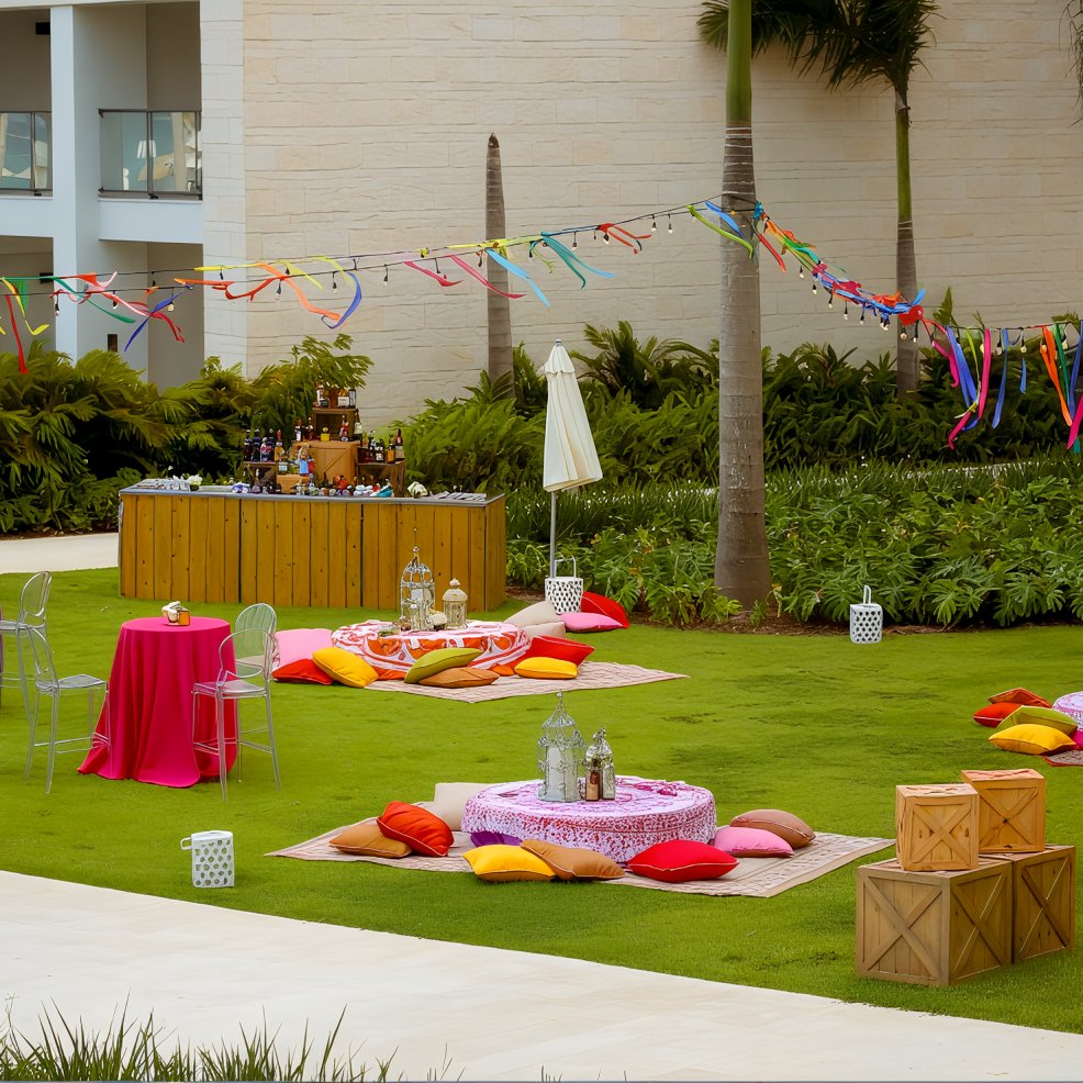 Destination wedding party scene with low tables, cushions, crates, and vibrant streamers on the lawn.