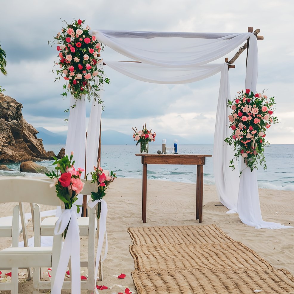 A destination wedding altar with white drapes and pink flowers on a sandy beach overlooking the sea.
