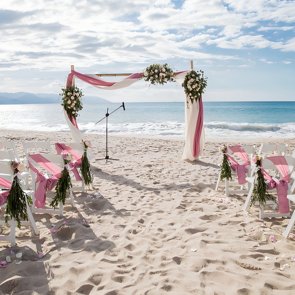 Destination wedding ceremony arranged on sandy beach with pink and white decor and floral arrangements.