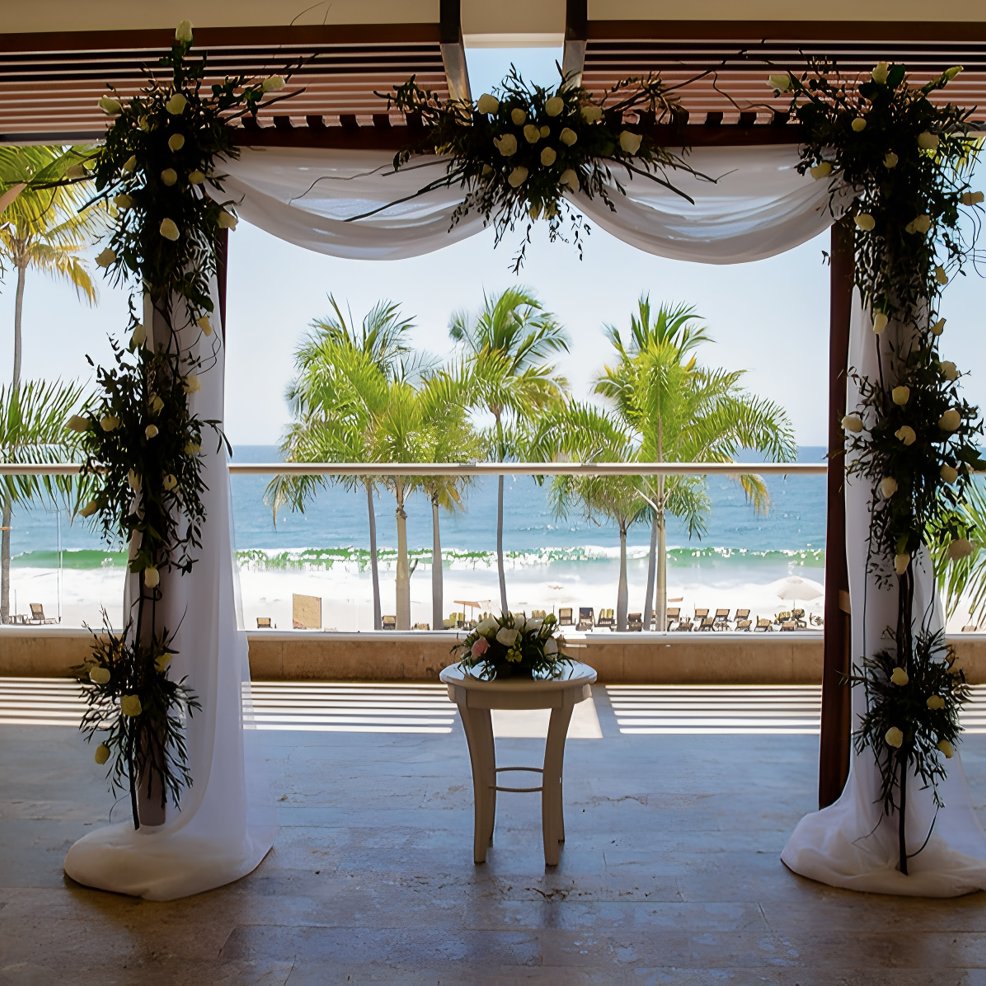 Destination wedding altar adorned with flowers, set above a sunny beach with palm trees in the background.