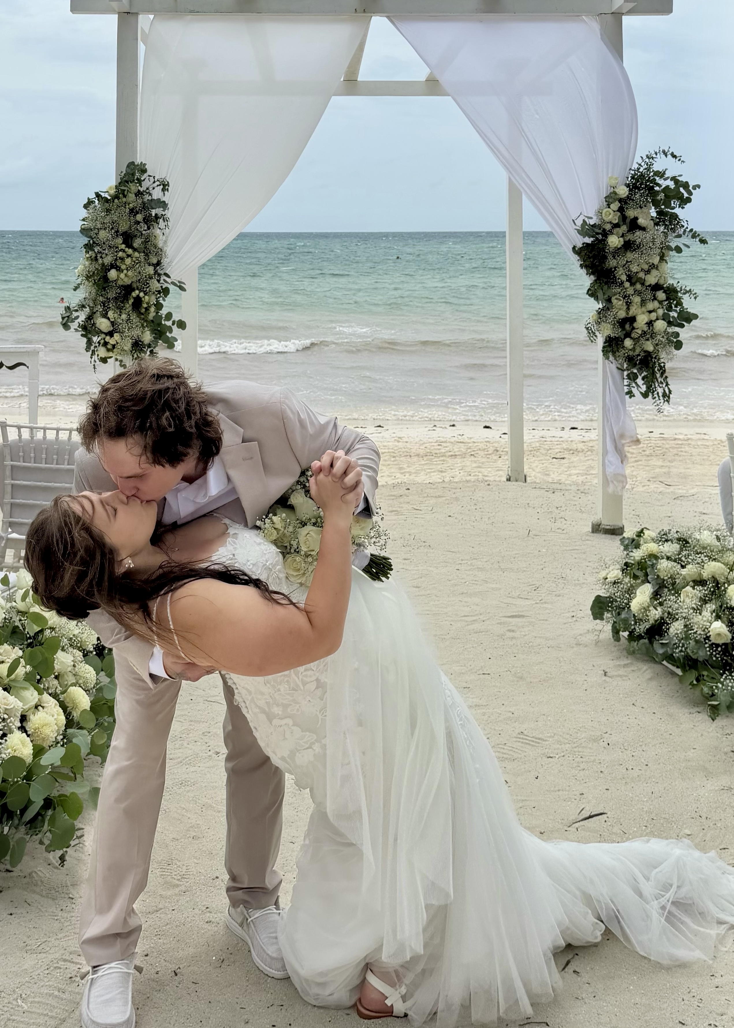 Stephanie and Connor kiss beneath a floral arch on the beach at their destination wedding.