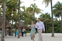 Stephanie and Connor stroll on a sandy beach lined with palm trees during their destination wedding.