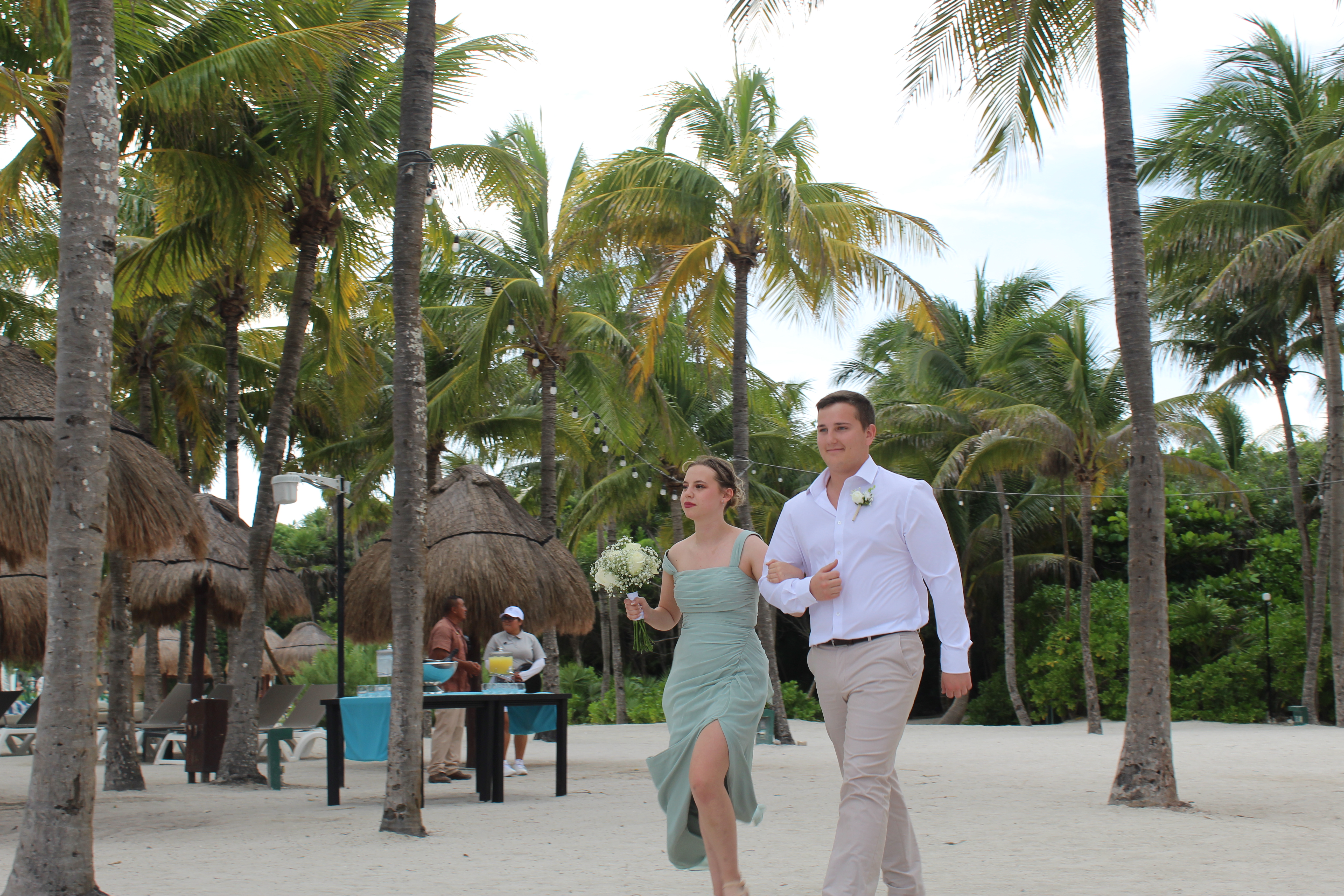 Stephanie and Connor stroll on a sandy beach lined with palm trees during their destination wedding.