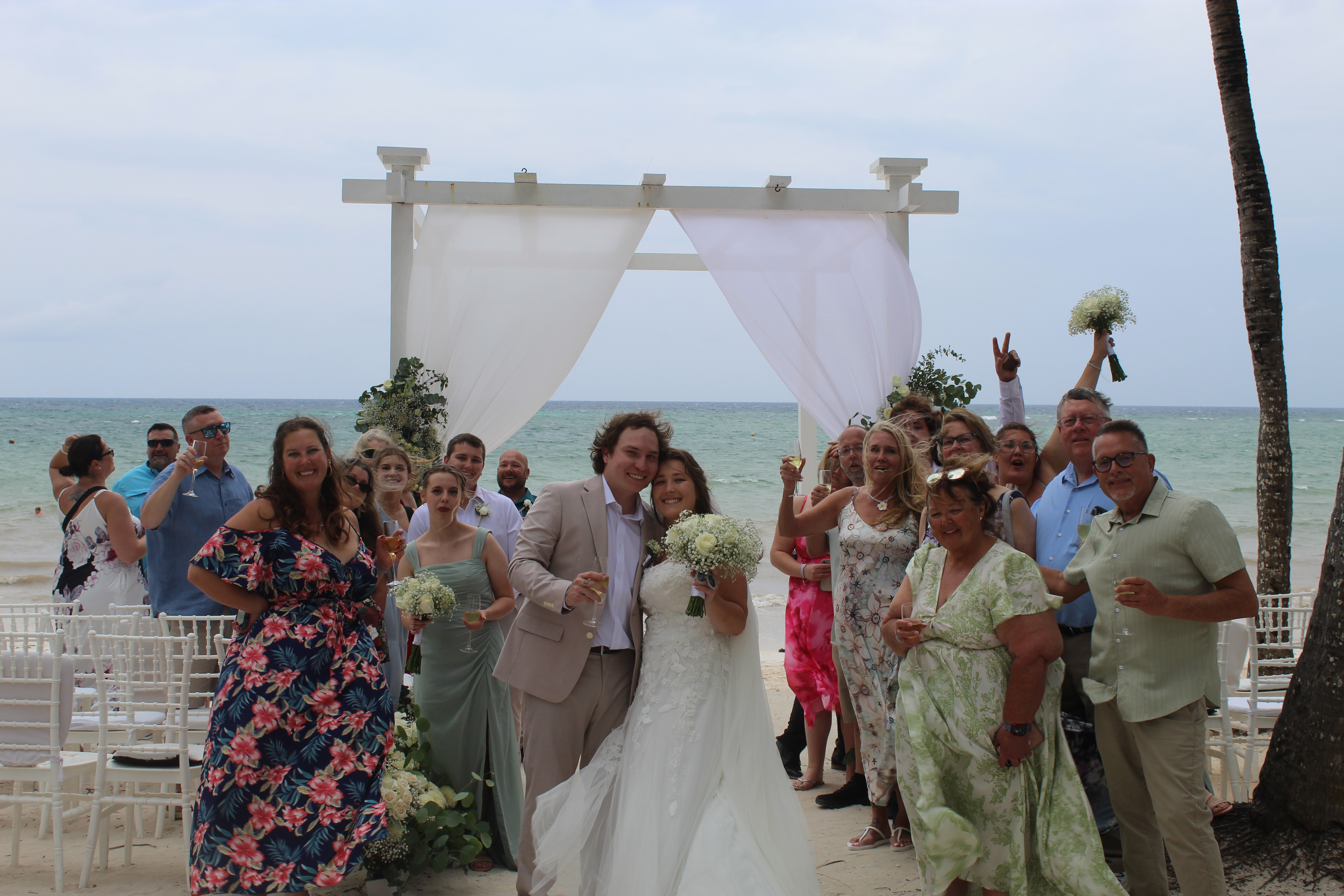Stephanie and Connor pose with guests at their destination wedding by a beachside altar under cloudy skies.