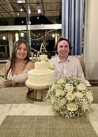 At their destination wedding, Stephanie and Connor sit at a table with a white cake and white roses.