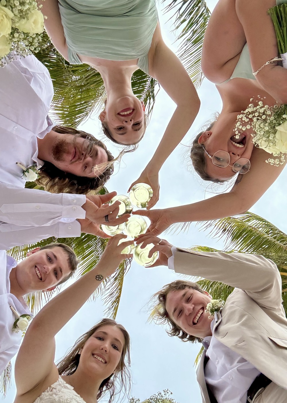 Six people in wedding attire toast drinks above, viewed from below at Stephanie and Connor&rsquo;s destination wedding.