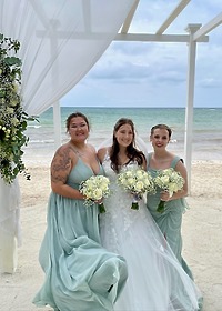 Three women in formal dresses hold bouquets on a beach at a destination wedding for Stephanie and Connor.