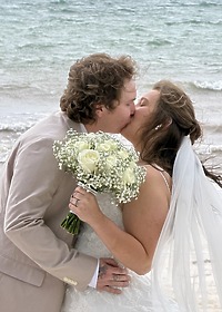 Stephanie and Connor share a kiss on a beach at their destination wedding, holding white roses and baby's breath.
