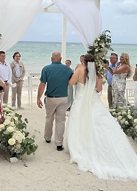 At a destination wedding, a bride walks down the sandy aisle with a man beside her at the beach.