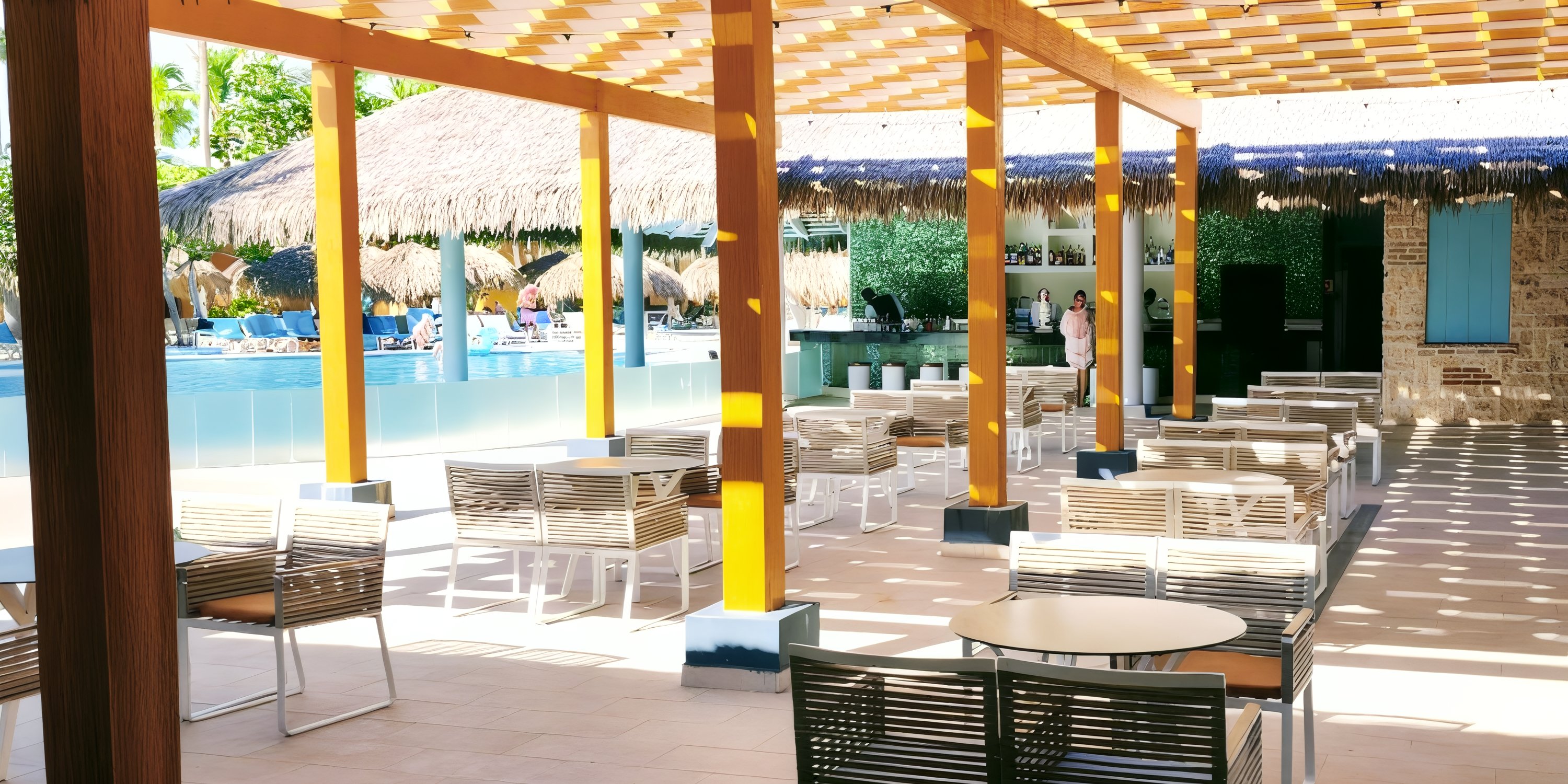 Poolside seating area with tables and chairs under a shaded pergola, ideal for a destination wedding near a bar.