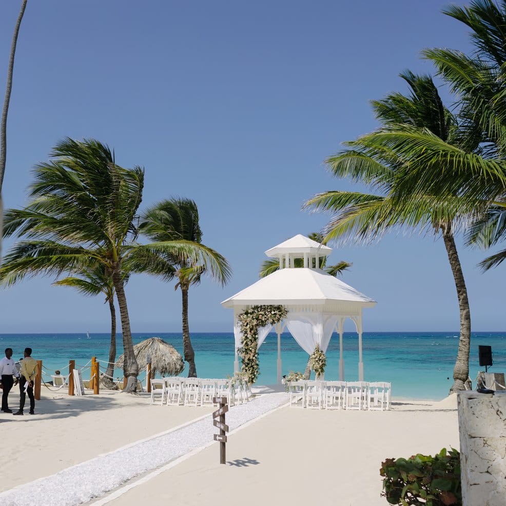 White gazebo prepared for a destination wedding, framed by palm trees and ocean in the background.