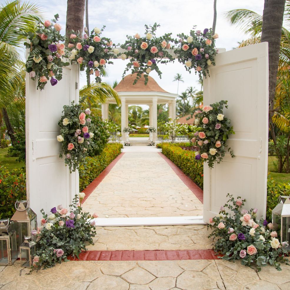 Flower-adorned white doors frame a stone walkway to a gazebo, perfect for a romantic destination wedding.