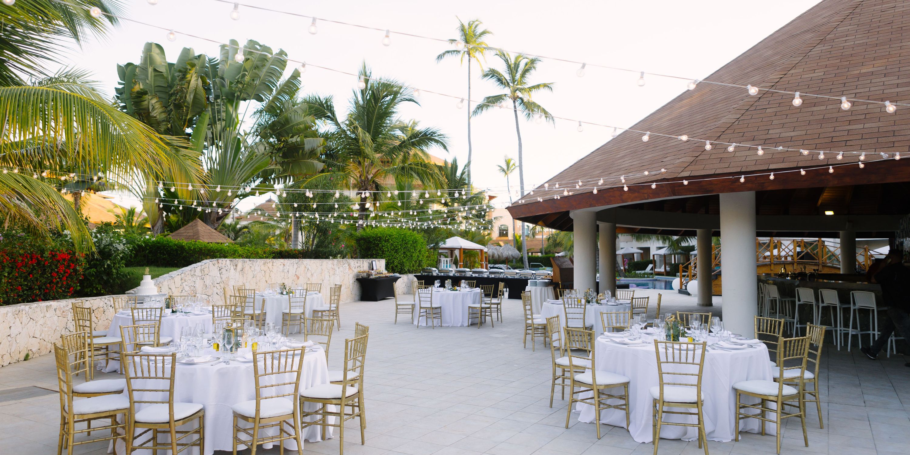 Outdoor patio set for a destination wedding, featuring round tables, chairs, string lights, and tropical plants.