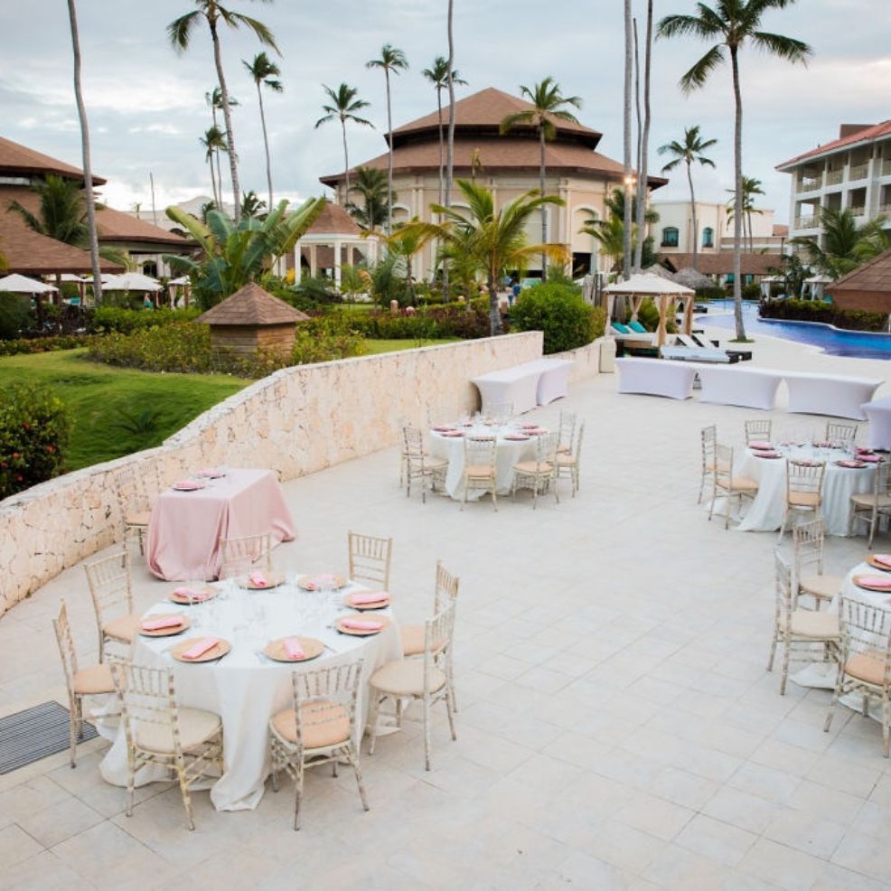 Outdoor wedding venue featuring round dining tables beside a pool with scenic resort buildings nearby.