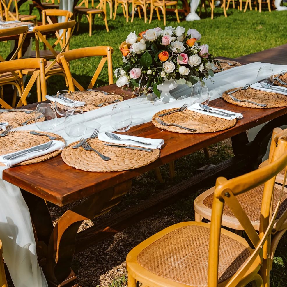 Outdoor wooden table arranged for a destination wedding, with woven placemats, glassware, and florals.