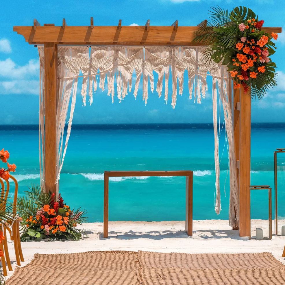 A wooden wedding arch adorned with flowers is set on a sandy beach for a destination wedding by turquoise water.