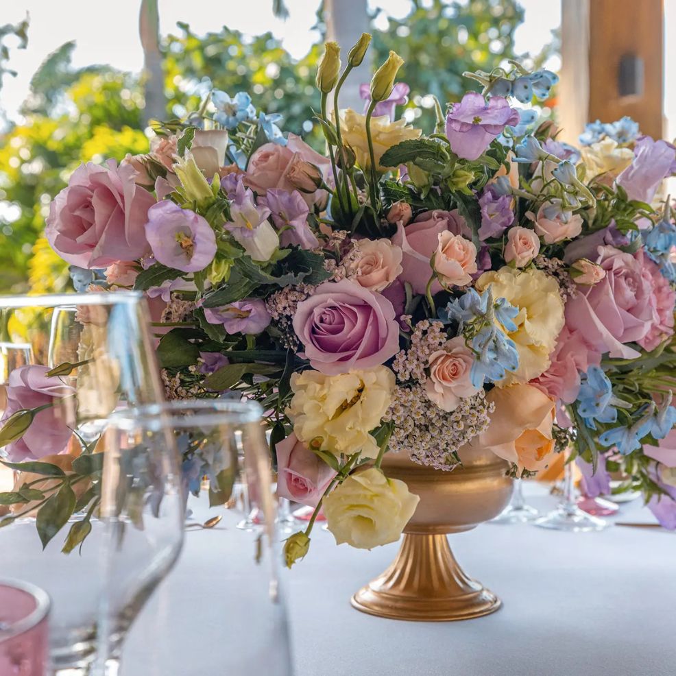 A gold vase with pastel flowers sits on a table, flanked by glassware at a destination wedding.
