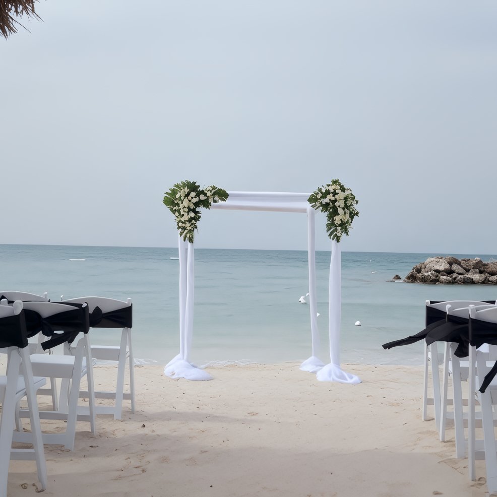 A wedding arch adorned with flowers stands on a sandy beach by the sea, surrounded by chairs for a destination wedding.