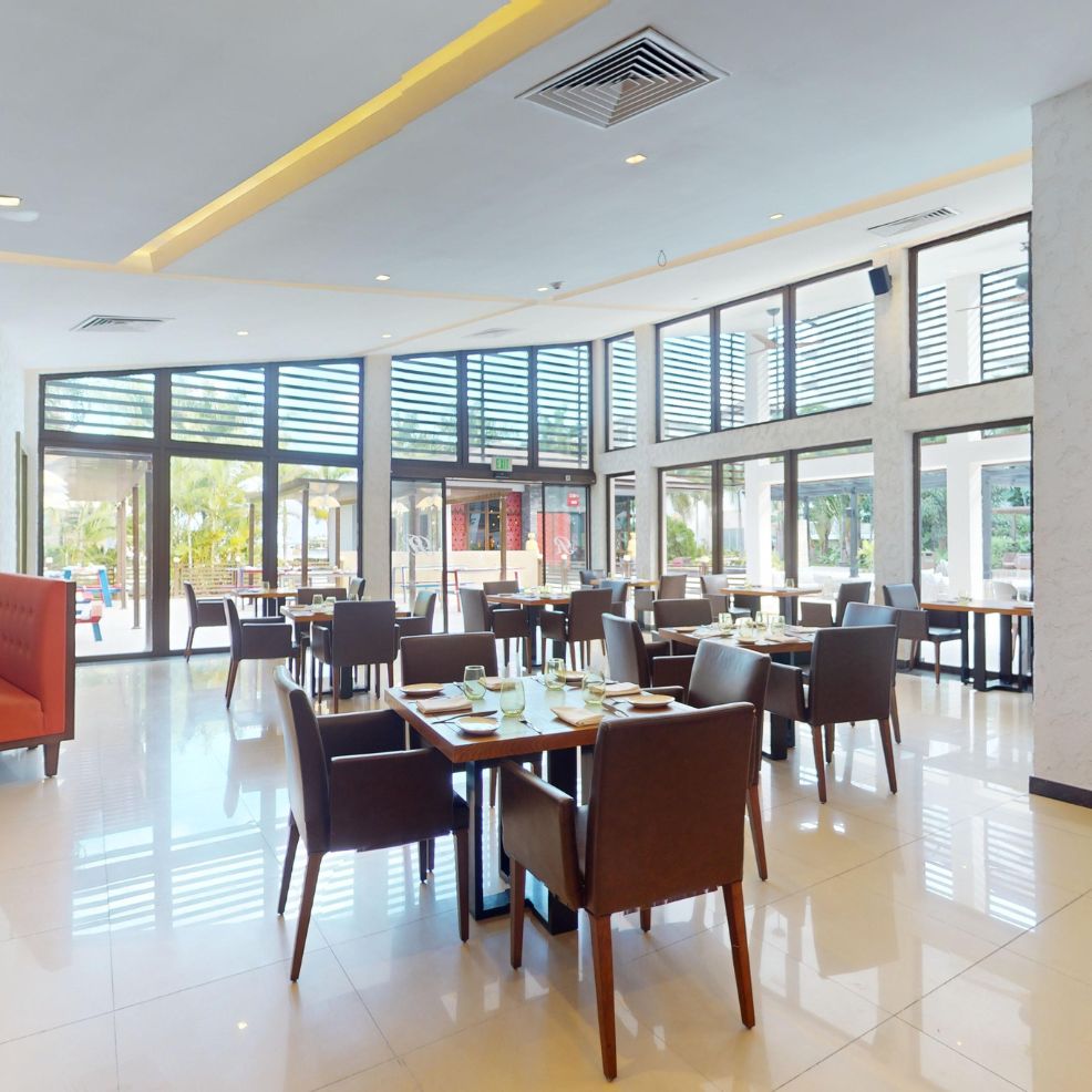 Modern restaurant set for a destination wedding, with empty tables, brown chairs, and sunlit large windows.