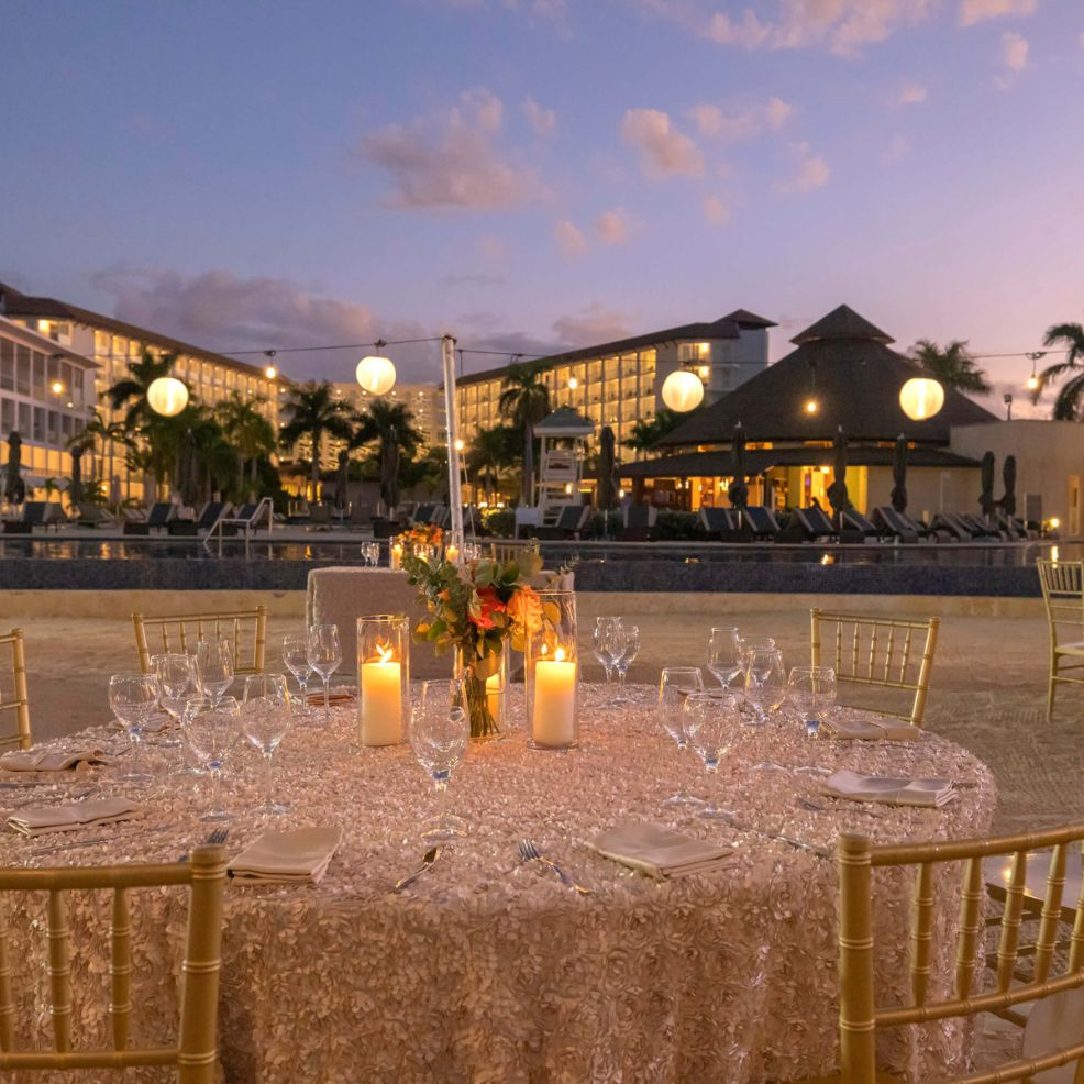 Round table with candles and glasses set up for a destination wedding by a pool at dusk near a resort.