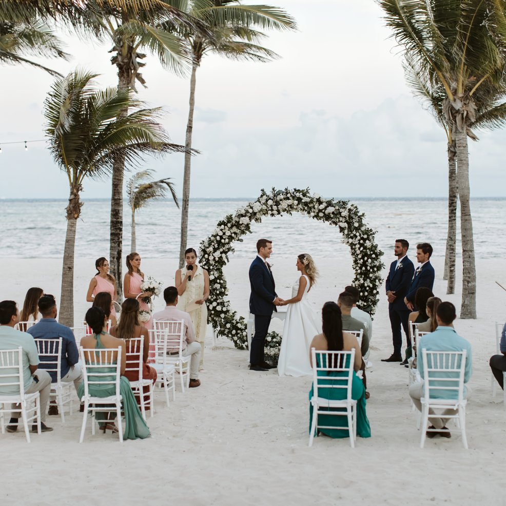 A couple stands at a destination wedding ceremony on the beach, guests seated and palm trees nearby.