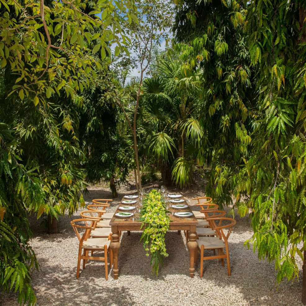 A wooden outdoor dining table set for a destination wedding, with plates and chairs amid lush greenery.