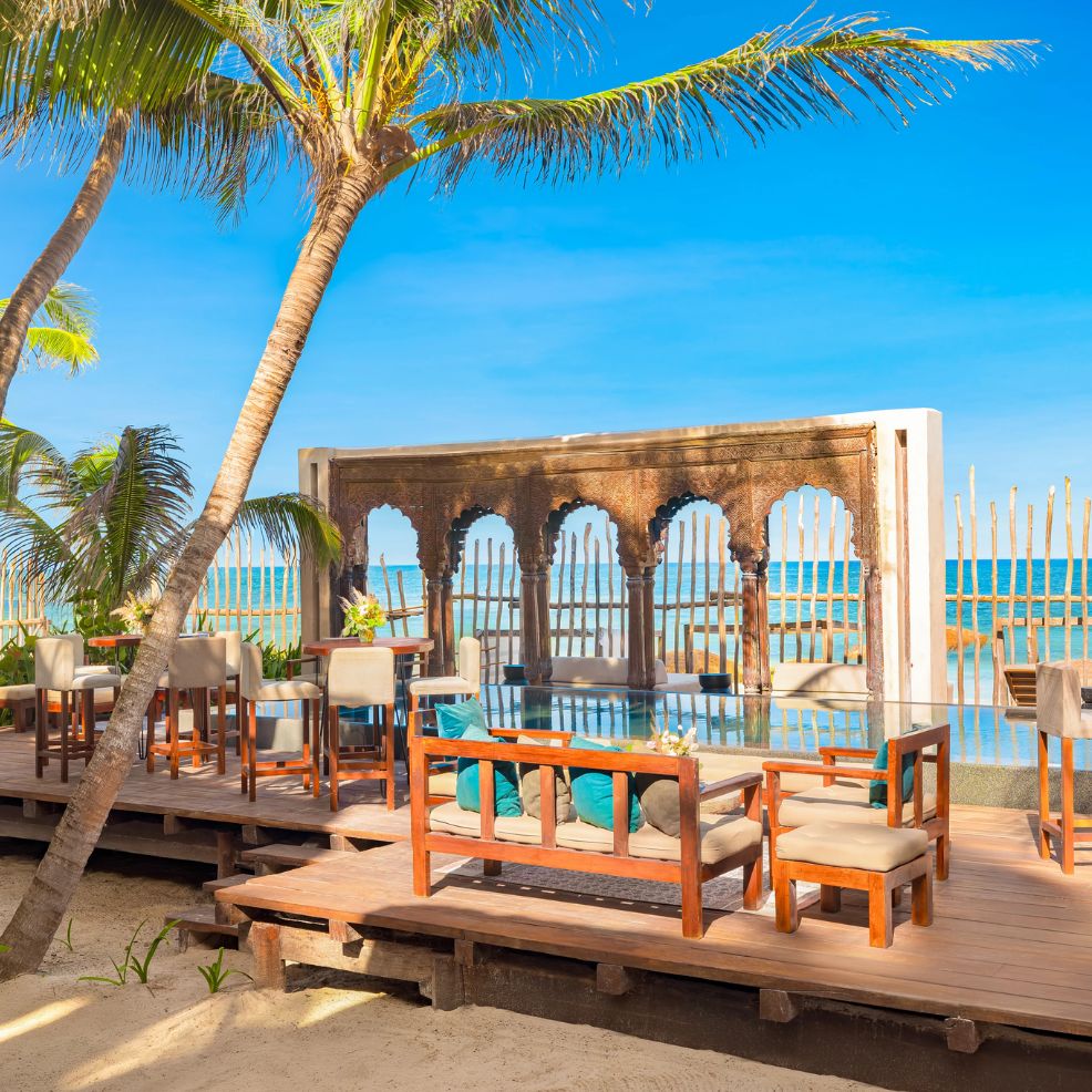Outdoor wooden seating area with palm trees, pool, and arched structure, perfect for a destination wedding.