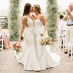 At their destination wedding, two brides share a kiss down the aisle as guests cheer and applaud.