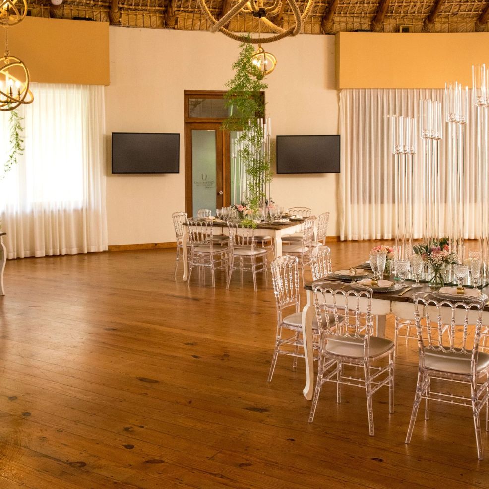 Round tables with clear chairs arranged for a destination wedding in a spacious, wood-floored room.