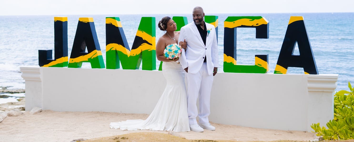 Bride and groom pose by a big "JAMAICA" sign at their oceanfront destination wedding.