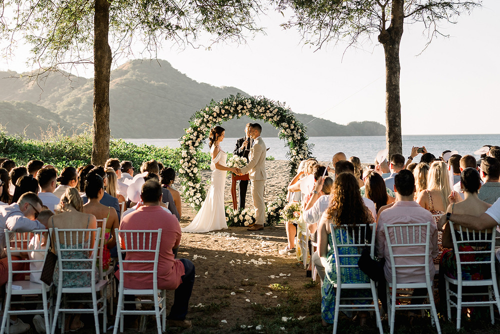 Destination wedding on a sandy beach, guests seated as the couple stands beneath a floral archway.