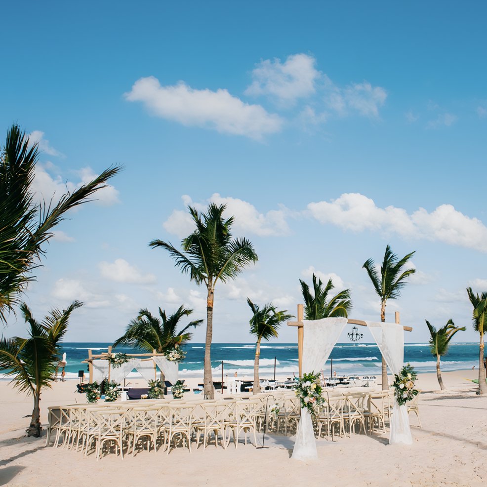 Destination wedding setup with chairs and a floral arch on a sandy beach, palm trees, and ocean behind.