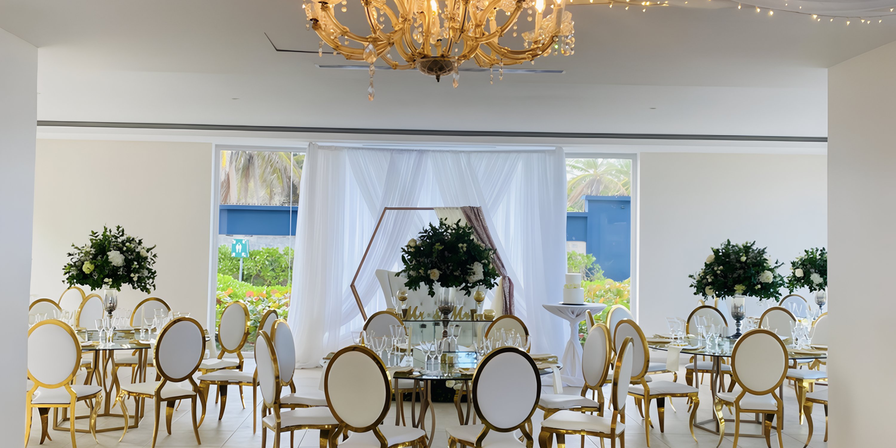 Banquet hall set for a destination wedding with round tables, gold chairs, flowers, and chandeliers.