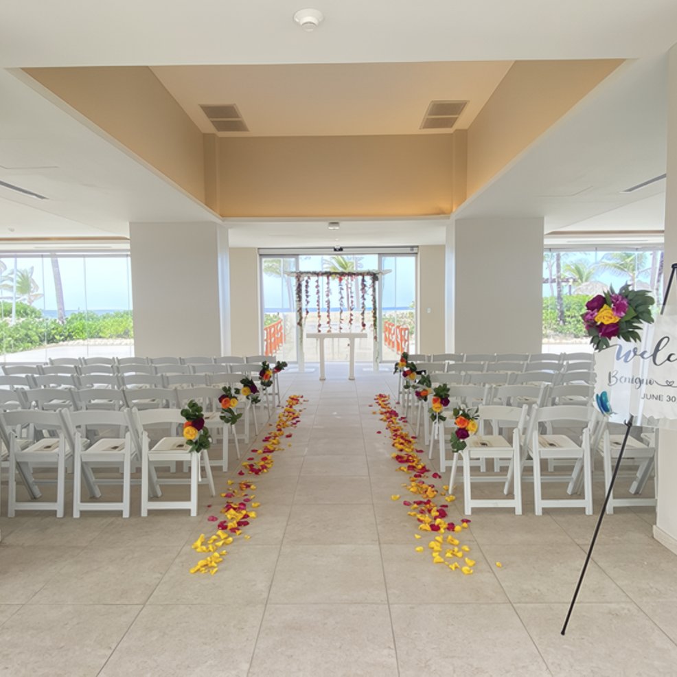 Elegant indoor destination wedding setup featuring white chairs, floral aisle accents, and a front altar.
