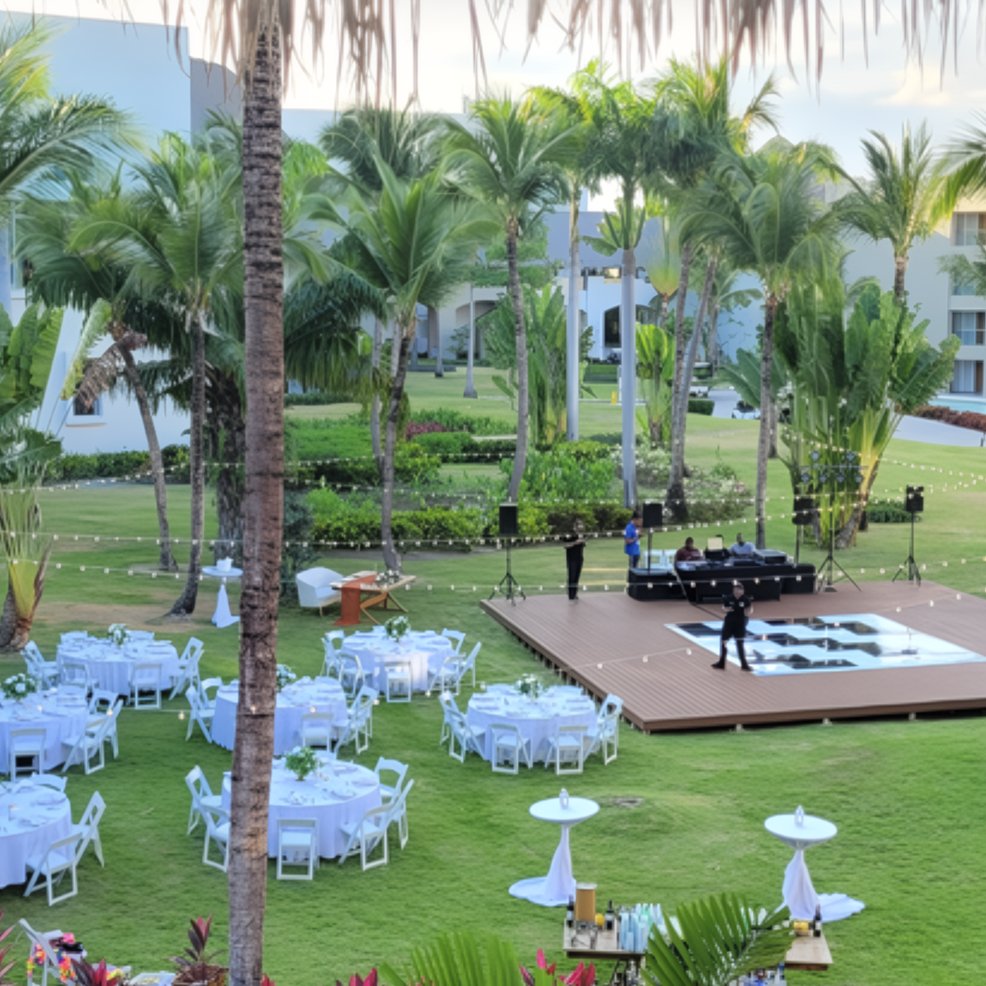 Destination wedding setup featuring round tables, chairs, and a stage on a lawn bordered by palm trees.