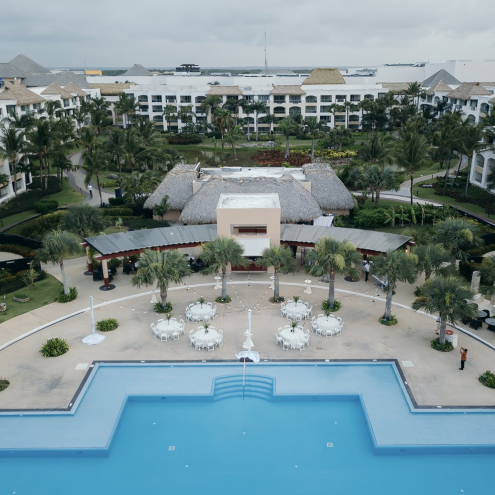 A large outdoor pool at a resort complex, with round tables set for a destination wedding under cloudy skies.