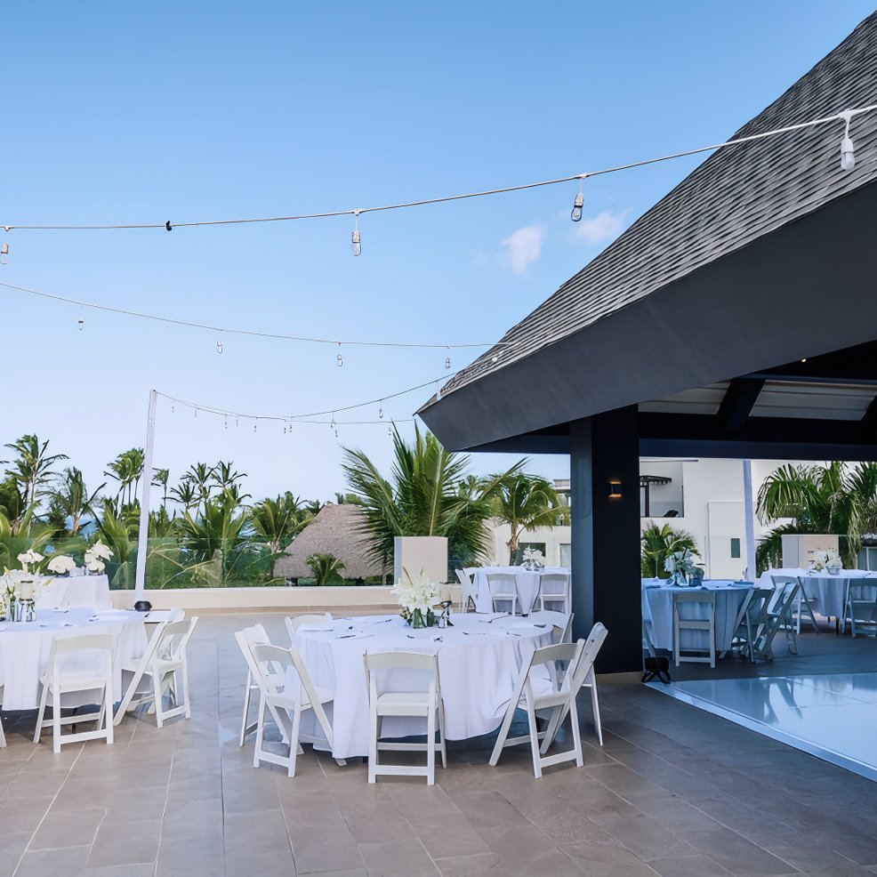 Outdoor patio arranged for a destination wedding, with round tables, white chairs, and glowing string lights.