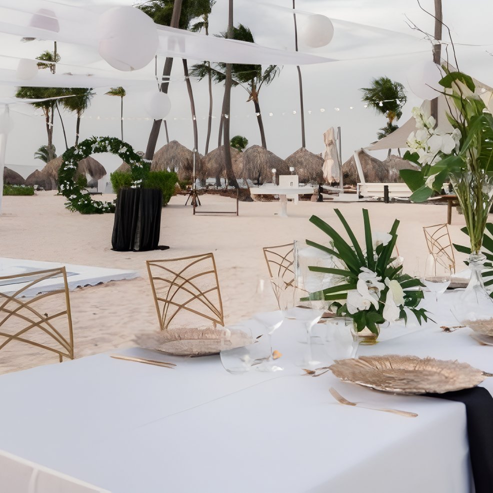 Destination wedding table set elegantly on sandy beach, featuring white decor and tropical palm trees.