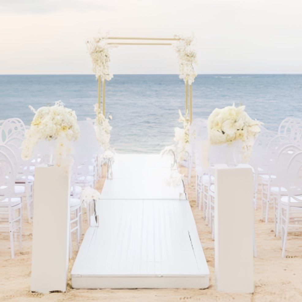 Beach destination wedding setup with a white arch, aisle, rows of chairs, and lush floral arrangements.