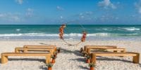 Wooden benches line a sandy beach, facing a floral arch for a destination wedding by the ocean.